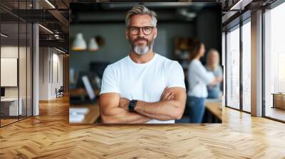 A confident businessman with gray hair and beard wears casual glasses and a watch, standing in a modern office environment with arms crossed and colleagues nearby. Wall mural