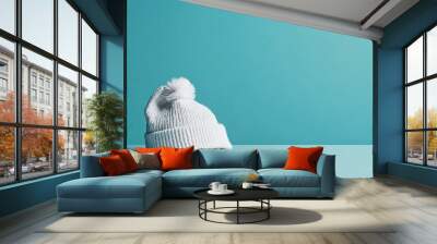 A cozy white knit hat with a fluffy pom-pom is displayed against a solid turquoise background, highlighting its texture and softness, suitable for winter fashion and warmth. Wall mural