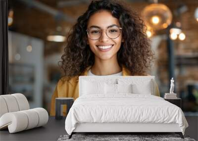 A joyful woman with curly hair and glasses is pictured holding her smartphone, standing in a stylish space with warm lighting and an inviting atmosphere. Wall mural