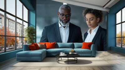 A mature man and a young woman examine information together on a tablet. The image reflects collaboration, mentorship, and technology in a corporate setting. Wall mural