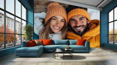 A smiling couple dressed in warm winter clothing, enjoying the snow, showcasing happiness and togetherness against a snowy, rustic background with wooden elements. Wall mural