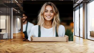 A woman wearing casual attire and an apron smiles while holding a wooden crate outdoors in a farm setting, implying freshness and enjoyment of rural life. Wall mural