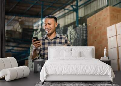 asian man factory employee removes safety helmet while using cell phone in warehouse Wall mural