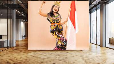 asian woman in traditional clothes of dayak tribe standing with clenched arm and bend her leg while holding the indonesian flag on isolated background Wall mural