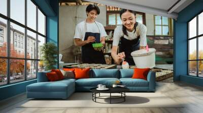 asian woman smiles wearing an apron while pouring the cake batter on the cake tin on the table in the background man wearing an apron helps to bake the cake. Wall mural