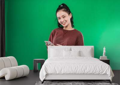 Portrait beautiful young Asian girl smiling cute holding a phone while looking at the camera isolated on a green background Wall mural