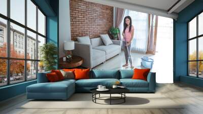 Teenage girl with smile holding a mop stick doing is mopping the living room on curtain windows background Wall mural