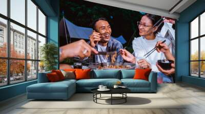 the guy in plaid shirt and woman in white knit sweater laughing together while eating the grilled beef from the grilled pan using the chopsticks Wall mural