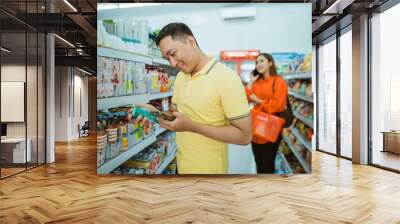 young man holding goods and mobile phone while standing shopping at minimarket Wall mural