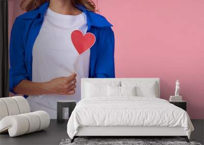 A young woman in a blue shirt holds a red paper heart in front of her chest. Pink studio background. The concept of women's health and diseases of the cardiovascular system Wall mural