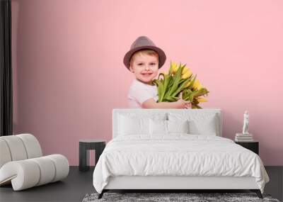 Adorable smiling child with spring flower bouquet looking at camera isolated on pink. Little toddler boy in hat holding yellow tulips as gift for mom. Copy space for text  Wall mural