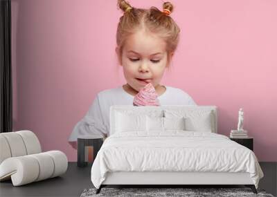 Cute child girl with pleasure eats tasty ice-cream on a pink studio background. Licks with closed eyes. The concept of baby food and a happy childhood Wall mural