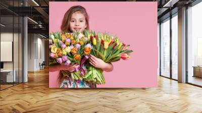 Cute smiling child girl holding bouquet of spring flowers tulips isolated on pink background. Little toddler girl gives a bouquet to mom. Copy space for text. Wall mural