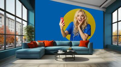 Image of young happy woman standing over a blue background. Girl peep out of a hole in the wall. Looking to the side to take a selfie on the mobile phone. Wall mural