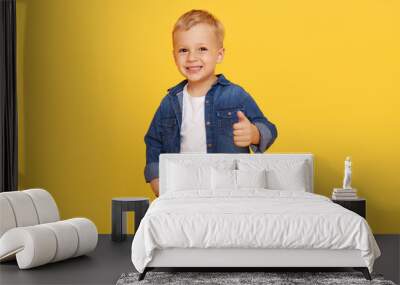 Joyful little child boy in denim clothes shows thumb up, OK symbol on yellow background. Advertising of childrens products and happy childhood Wall mural