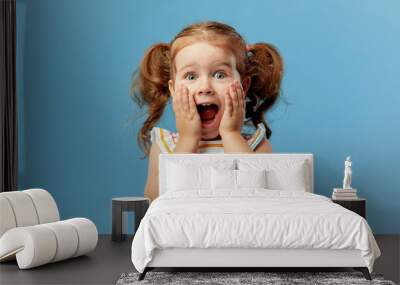 Portrait of surprised cute little toddler girl child standing isolated over blue background. Looking at camera. hands near open mouth Wall mural