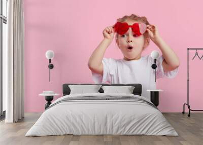 Portrait of surprised cute little toddler girl in the heart shape sunglasses. Child with open mouth having fun isolated over pink background. Looking at camera. Wow funny face Wall mural