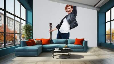 Young woman musician in a black leather jacket with an acoustic guitar in hand on a gray background. He laughs and plays rock and roll loudly. Full-length portrait Wall mural