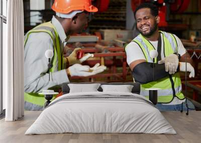 African factory worker or engineer wearing bandages splint. pain in elbow ache in arm from hard work and coworker helping him in the factory Wall mural