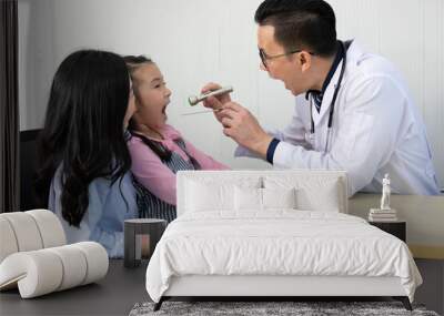 asian doctor examining to little girl mouth with tongue depressor and mother by her side Wall mural