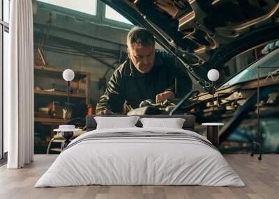 In the bright workshop of a car repair shop, an auto mechanic with a scope in his hands, works on the open bonnet of a car. Blur effect in the background Wall mural