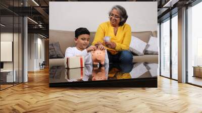 Indian/asian Grandma teaching her grand son importance of savings, putting coins into pink piggy bank. Wall mural