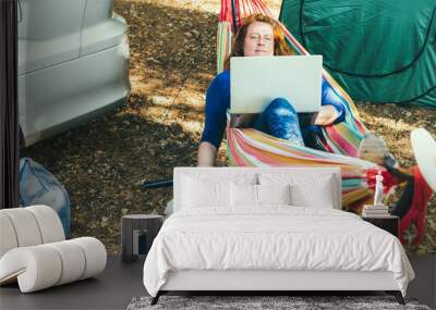 Adult woman working on laptop pc while lying in hammock, stroking cockapoo puppy near motorhome on camping trip. Female living on camper car with animals and travel the world. Remote worker people. Wall mural