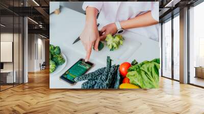 Go vegan. Close up woman cutting broccoli while cooking healthy dish according to the active online mobile application with Vegan diet program on her phone. Healthy lifestyle, weight loss concept Wall mural