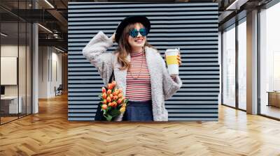 Happy, smiling young fashion woman in heart shaped glasses with reusable coffee cup and bouquet of tulip flowers on gray striped wall background. Eco friendly urban city street fashion. Spring mood Wall mural