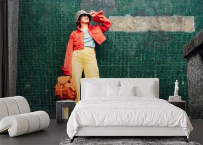 Hipster young woman in bright clothes, sun glasses, backpack bag and bucket hat posing on the green tile wall background. Urban city street fashion. Fashion blogger. Selective focus. Copy space Wall mural