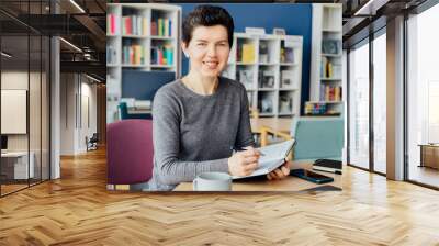 Smiling neutral gender middle-aged woman making notes in paper notebook, using laptop, working in open space office. Adult students studing, making research in public library. Academic research. Wall mural