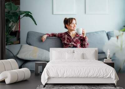 Young smiling woman sitting on sofa and looking side up while drinking coffee or tea. Young brunette woman relaxing after housekeeping, home cleaning. Portrait of relaxed female resting at home Wall mural