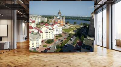 An aerial view of a cityscape featuring colorful buildings with red rooftops and a striking church with golden domes near a river. The tranquil water and distant skyline create a beautiful urban view Wall mural