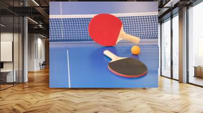 Orange ball for table tennis and two rackets of red and black color on a blue table with a grid Wall mural