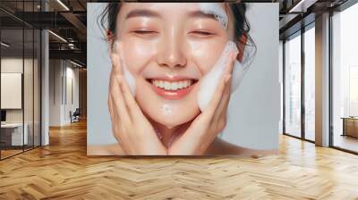 Close-up portrait of smiling  asian woman washing face with soap foam, isolated on light grey background Wall mural