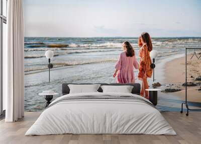 Two teen caucasian barefooted girls from behind wearing airy dresses holding their hands and walking on the beach in sunny summer day. Sisterhood and friendship Wall mural