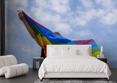 Bisexual, girl, lesbian, woman, transgender, homosexsual holding in hand a rainbow LGBT gender identity flag on sky background with clouds on a sunny day and celebrating a gay parade in pride month Wall mural