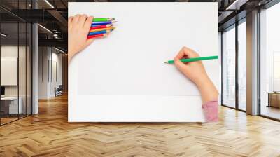 Top view of little child's hand drawing by green pencil on white paper holding bunch of colored pencils in her hand. Selective focus. Wall mural