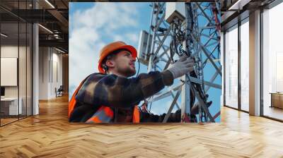 Helmeted male engineer works in the field with a telecommunication tower that controls cellular electrical installations to inspect and maintain 5G networks installed on high-rise buildings Wall mural