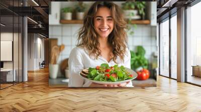 Cute, young girl in kitchen holding plate with salad of fresh vegetables, herbs and smiling Wall mural