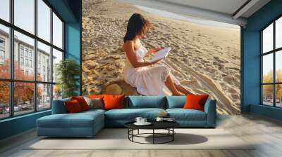 Romantic girl in a white dress reads a book sitting on a stone on the beach. View from above. Wall mural