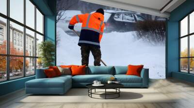 Man worker in orange uniform with a shovel cleaning the street near the car parking. Snow removal in winter city during snowstorm Wall mural