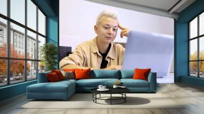 Smiling girl with short blonde hair sitting at office table with laptop. Tomboy lifestyle, concept of inspiration at work and creativity Wall mural