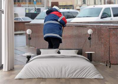 Woman worker in uniform cleaning a city street on cars background. Janitor washing sidewalk in winter Wall mural