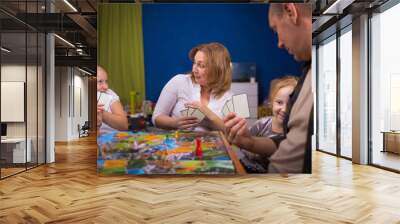 Board game concept. The family is having fun playing together at home in a board game. Board game field, many figures, coins and sandglass. Four people play, holding cards joyful emotions on the face. Wall mural