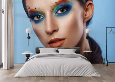 Fashion creative make up. Beautiful portrait of young woman with clean fresh skin and bright make up with rhinestones on her face posing on a bright blue background Wall mural