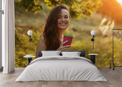 Young beautiful smiling woman in park and holding paper cup with coffee or tea on a tree and bush background in golden hour free space for text Wall mural