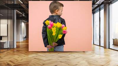 Boy hiding flowers of yellow and pink tulips behind itself, isolated on light orange background. Copy space Wall mural