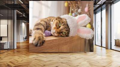 Cute kitty looks at the camera in a bunny costume. The cat is lying on a wooden background wearing a cute hat with bunny ears. Happy Easter concept Wall mural