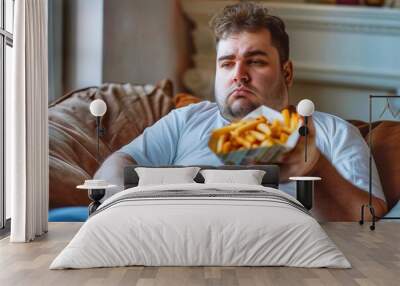 Fat man eating french fries sitting on a sofa in the living room at home Wall mural